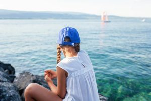 Une jeune fille portant une casquette de baseball bleue et une robe blanche profite d'un moment de sérénité au bord du lac, regardant les voiliers glisser sur l'eau.